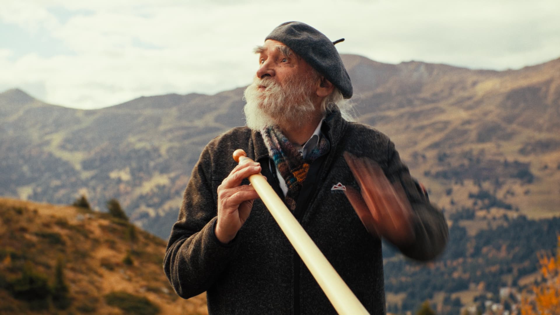 Älterer Mann mit Baskenmütze und Alphorn vor herbstlichem Bergpanorama