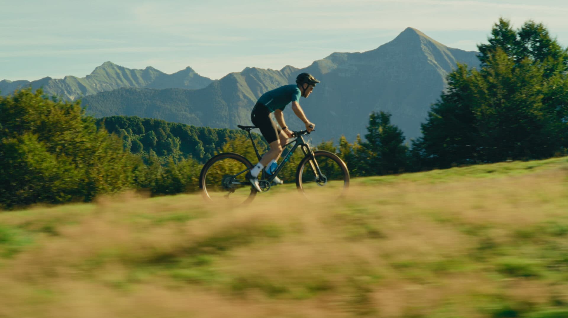 Mountainbiker fährt über eine Wiese, Tessiner Berge im Hintergrund